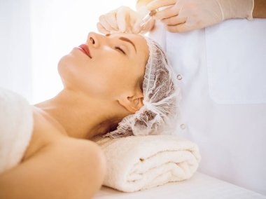 Güzel bir kadın güneşli tıp merkezinde kapalı gözlerle güzellik iğneleri yaptırıyor. Güzellik uzmanı elleri şırıngayla kadın yüzüne güzellik prosedürü uyguluyor. Kozmetik tıp ve cerrahi konsepti