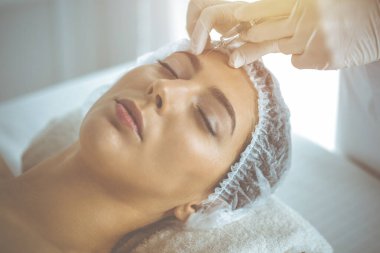 Güzellik uzmanı güneşli klinikte genç esmer kadının yüzüne şırıngayla güzellik prosedürü uyguluyor. Kozmetik ilaç ve ameliyat, güzellik iğneleri.