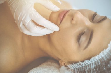 Güzellik uzmanı güneşli klinikte genç esmer kadının yüzüne şırıngayla güzellik prosedürü uyguluyor. Kozmetik ilaç ve ameliyat, güzellik iğneleri.