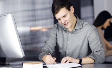 Yeşil gömlekli genç işadamı ve programcı iş planına bazı notlar yazarken, arka planda bayan meslektaşıyla birlikte bir dolapta oturuyor.