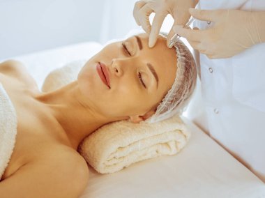 Güzel bir kadın tıp merkezinde kapalı gözlerle güzellik iğneleri yaptırıyor. Güzellik uzmanı elleri şırıngayla kadın yüzüne güzellik prosedürü uyguluyor.