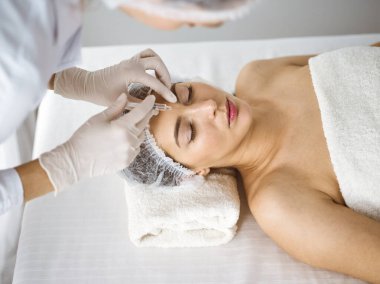 Güzellik uzmanı güneşli klinikte genç esmer kadının yüzüne şırıngayla güzellik prosedürü uyguluyor. Kozmetik ilaç ve ameliyat, güzellik iğneleri.
