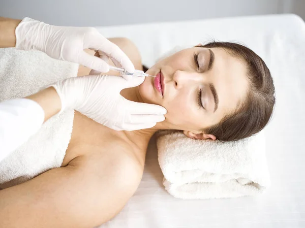 Beautician doing beauty procedure with syringe to face of young brunette woman in sunny clinic. Cosmetic medicine and surgery, beauty injections