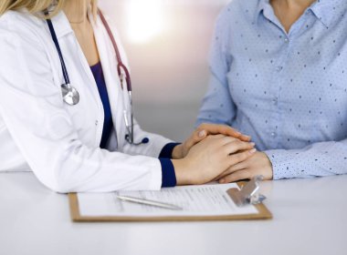 Bilinmeyen kadın doktor, bir klinikteki güneşli dolapta yan yana otururken hastasına güven vermek için elini tutuyor. Kadın hekim bir pano ve steteskop kullanıyor.