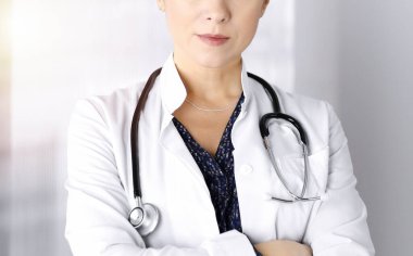 Gülümseyen profesyonel kadın doktor, güneşli bir klinikte kollarını kavuşturmuş duruyor. Steteskopu olan dost canlısı bir kadın doktorun portresi. Hastanede mükemmel bir tıbbi hizmet.