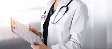 Neşeli, zeki bir kadın doktor güneşli bir klinikte dururken elinde bir pano tutuyor. Dost canlısı, steteskoplu bir kadın doktorun portresi. Mükemmel bir tıbbi hizmet.