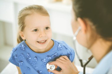 Bir çocuğu steteskopla muayene eden bir doktor. Mavi elbiseli mutlu gülümseyen kız hasta her zamanki sağlık kontrolünde. Tıp konsepti