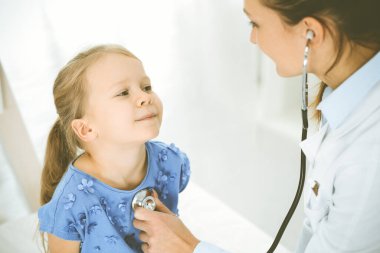 Bir çocuğu steteskopla muayene eden bir doktor. Mavi elbiseli mutlu gülümseyen kız hasta her zamanki sağlık kontrolünde. Tıp konsepti