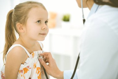 Bir çocuğu steteskopla muayene eden bir doktor. Parlak renkli elbise giymiş mutlu gülümseyen kız hasta her zamanki sağlık kontrolünde.