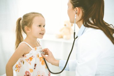 Bir çocuğu steteskopla muayene eden bir doktor. Parlak renkli elbise giymiş mutlu gülümseyen kız hasta her zamanki sağlık kontrolünde.