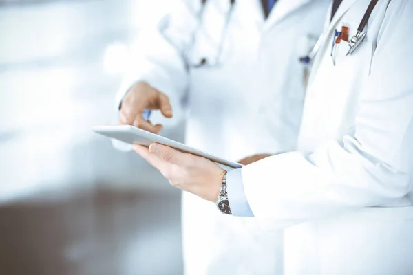 Two unknown doctors with stethoscopes discussing medical exam resoults, standing at hospital office. Physicians use a computer tablet for filling up medication names records. Perfect medical service