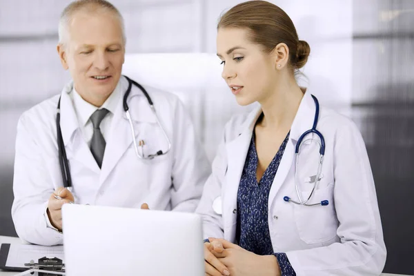 Elderly doctor and his female colleague discussing current disease therapy while sitting at working place in clinic. Teamwork in hospital. Medicine and healthcare concept