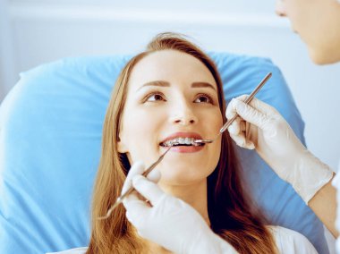 Diş hekiminde dişçi tarafından muayene edilen diş telleriyle gülümseyen genç bir kadın. Sağlıklı dişler ve tıbbi bakım konsepti