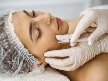 Güzellik uzmanı güneşli klinikte genç esmer kadının yüzüne şırıngayla güzellik prosedürü uyguluyor. Kozmetik ilaç ve ameliyat, güzellik iğneleri.