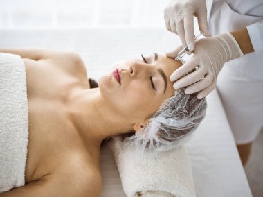 Güzellik uzmanı güneşli klinikte genç esmer kadının yüzüne şırıngayla güzellik prosedürü uyguluyor. Kozmetik ilaç ve ameliyat, güzellik iğneleri.