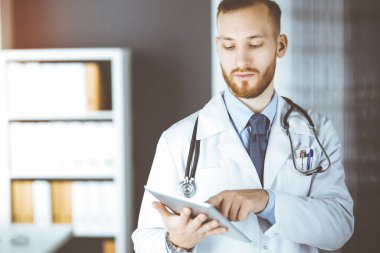 Güneşli klinikte tablet bilgisayar kullanan arkadaş canlısı kızıl sakallı doktor. Tıp konsepti