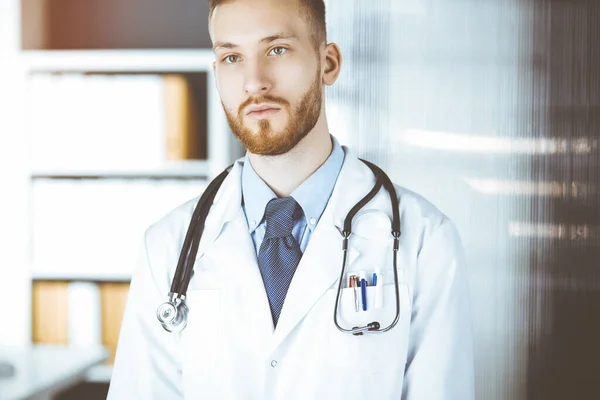 Red-bearded doctor standing straight in sunny clinic near his working place. Medicine concept