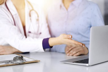 Bilinmeyen bir kadın, doktor ve hastası bir klinikteki güneşli dolapta birlikte otururken hastaların kan testlerini tartışıyorlar. Steteskoplu bir kadın doktor dizüstü bilgisayar kullanıyor.