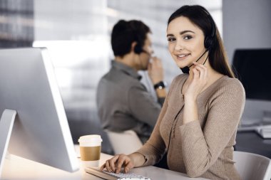 Modern bir ofiste meslektaşıyla birlikte masa başında otururken, kulaklıklı genç bir kız bir firma müşterisiyle konuşuyor. İş yerindeki çağrı merkezi operatörleri