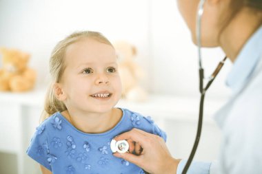 Bir çocuğu steteskopla muayene eden bir doktor. Mavi elbiseli mutlu gülümseyen kız hasta her zamanki sağlık kontrolünde. Tıp konsepti