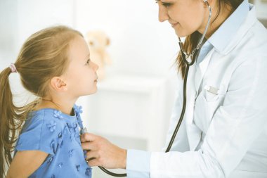 Bir çocuğu steteskopla muayene eden bir doktor. Mavi elbiseli mutlu gülümseyen kız hasta her zamanki sağlık kontrolünde. Tıp konsepti