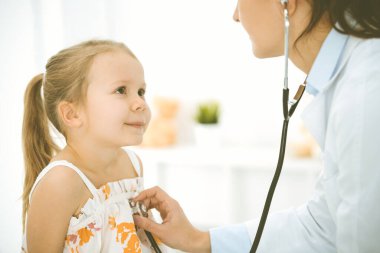 Bir çocuğu steteskopla muayene eden bir doktor. Parlak renkli elbise giymiş mutlu gülümseyen kız hasta her zamanki sağlık kontrolünde.