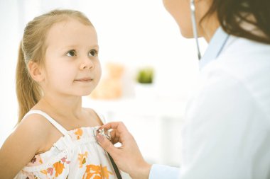 Bir çocuğu steteskopla muayene eden bir doktor. Parlak renkli elbise giymiş mutlu gülümseyen kız hasta her zamanki sağlık kontrolünde.