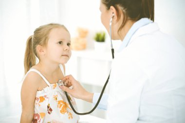 Bir çocuğu steteskopla muayene eden bir doktor. Parlak renkli elbise giymiş mutlu gülümseyen kız hasta her zamanki sağlık kontrolünde.