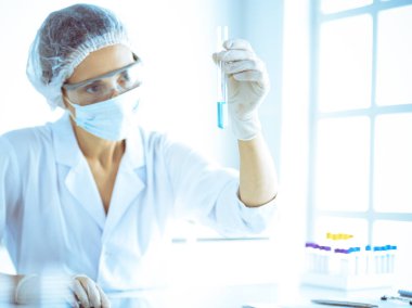 Koruyucu gözlüklü profesyonel kadın bilim adamı labaratuvarda mavi renkte reaksiyonlarla araştırma tüpü kullanıyor. Tıp ve bilim araştırmaları kavramları