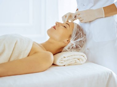 Güzel bir kadın tıp merkezinde kapalı gözlerle güzellik iğneleri yaptırıyor. Güzellik uzmanı elleri şırıngayla kadın yüzüne güzellik prosedürü uyguluyor.