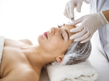 Güzellik uzmanı güneşli klinikte genç esmer kadının yüzüne şırıngayla güzellik prosedürü uyguluyor. Kozmetik ilaç ve ameliyat, güzellik iğneleri.