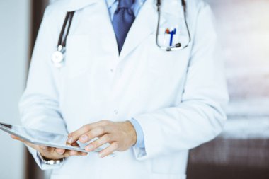 Dost canlısı sakallı doktor çalışma yerindeki güneşli klinikte tablet bilgisayar kullanıyor.
