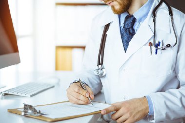 Dost canlısı kızıl sakallı doktor klinikte pano ile oturup yazıyor.