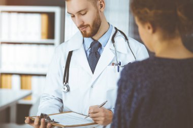 Dost canlısı kızıl sakallı doktor ve hasta kadın güneşli klinikte otururken sağlık muayenesini tartışıyorlar. İlaç, sağlık ve virüs koruma programları