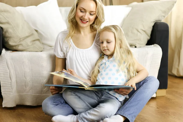 Happy family. Blonde young mother reading a book to her cute daughter while sitting at wooden floor. Motherhood concept