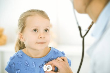 Bir çocuğu steteskopla muayene eden bir doktor. Mavi elbiseli mutlu gülümseyen kız hasta her zamanki sağlık kontrolünde. Tıp konsepti