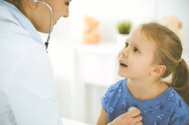 Bir çocuğu steteskopla muayene eden bir doktor. Mavi elbiseli mutlu gülümseyen kız hasta her zamanki sağlık kontrolünde. Tıp konsepti
