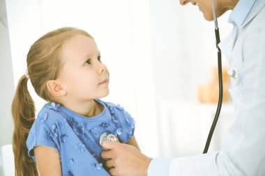 Bir çocuğu steteskopla muayene eden bir doktor. Mavi elbiseli mutlu gülümseyen kız hasta her zamanki sağlık kontrolünde. Tıp konsepti
