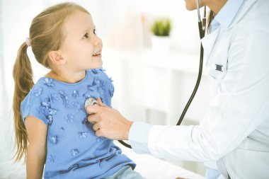 Bir çocuğu steteskopla muayene eden bir doktor. Mavi elbiseli mutlu gülümseyen kız hasta her zamanki sağlık kontrolünde. Tıp konsepti