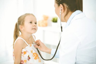 Bir çocuğu steteskopla muayene eden bir doktor. Parlak renkli elbise giymiş mutlu gülümseyen kız hasta her zamanki sağlık kontrolünde.