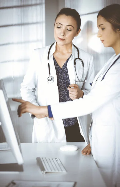 Two female physicians are discussing medical therapy, while standing at the table in a sunny clinic office. Doctors use pc computer at work. Teamwork in medicine