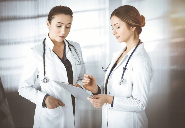 Two female physicians are discussing medical therapy, while standing at the table in a sunny clinic office. Doctors use pc computer at work. Teamwork in medicine