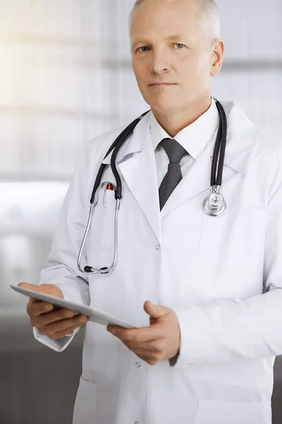 Elderly confident doctor standing straight and using tablet in sunny clinic. Perfect medical service concept