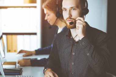 Kızıl sakallı iş adamı güneşli bir ofiste otururken kulaklıkla konuşuyor. Çağrı merkezinde çeşitli insanlar grubu