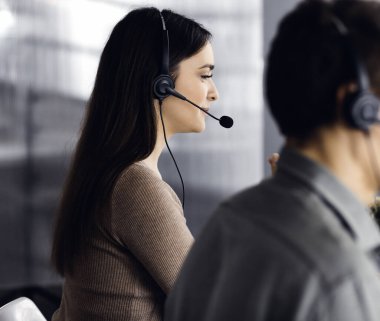 Kulaklık takmış iki genç bir ofiste masada otururken müşterilerle konuşuyor. Kadınlara odaklan. İş yerindeki çağrı merkezi operatörleri