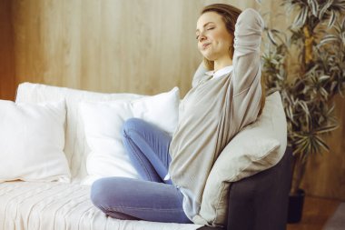 Mutlu esmer kadın ellerini başının arkasına koyuyor koltuğa yaslanıyor, Avrupalı kadın esniyor ve tembel haftasonunun keyfini çıkarıyor ya da sıcak oturma odasında tatilin tadını çıkarıyor.