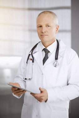 Yaşlı, kendine güveni tam bir doktor güneşli klinikte tablet kullanıyor. Mükemmel tıbbi servis kavramı
