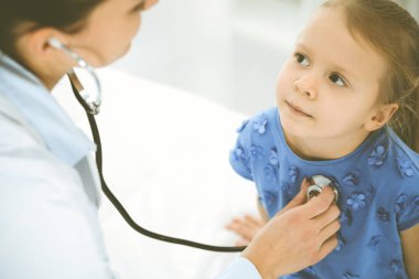Bir çocuğu steteskopla muayene eden bir doktor. Mavi elbiseli mutlu gülümseyen kız hasta her zamanki sağlık kontrolünde. Tıp konsepti