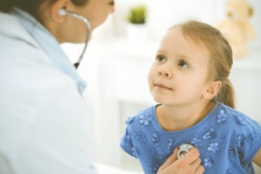 Bir çocuğu steteskopla muayene eden bir doktor. Mavi elbiseli mutlu gülümseyen kız hasta her zamanki sağlık kontrolünde. Tıp konsepti