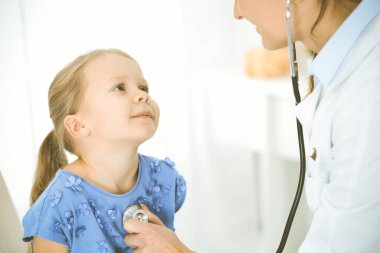 Bir çocuğu steteskopla muayene eden bir doktor. Mavi elbiseli mutlu gülümseyen kız hasta her zamanki sağlık kontrolünde. Tıp konsepti